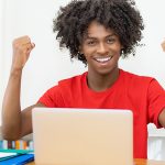 High school student sitting at laptop raising two fists in celebration