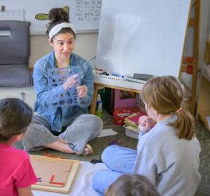 A Montessori teacher sits on the floor, guiding young students through a lesson.