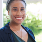 Professional headshot of an instructor wearing a navy blazer and green top, with a soft-focus outdoor background of greenery.