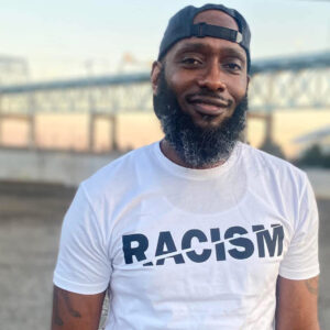 Kevin Martin stands outdoors near a bridge at sunset, wearing a white T-shirt with the word "RACISM" with a slash through it displayed across the front. Martin is a University of Delaware human services student whose educational journey was shaped by support from the ACCESS Center.