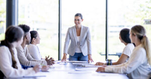 Person standing at the head of a conference table leading a meeting with three colleagues seated on each side.