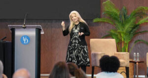Nancy Dibert delivers a presentation at a University of Delaware event, standing onstage beside a UD podium with audience members visible in the foreground.